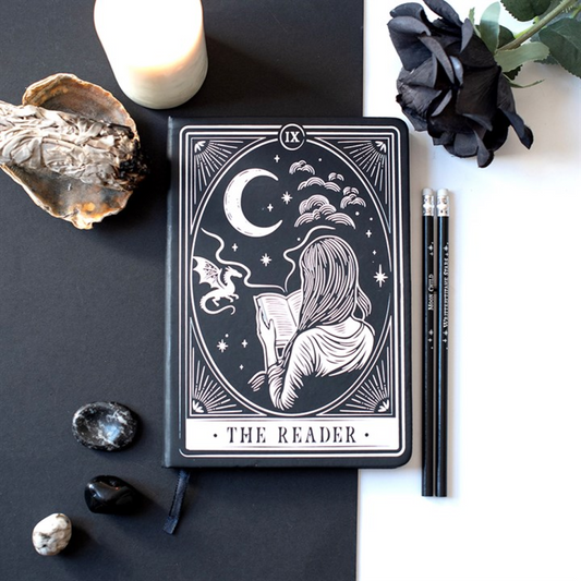 A tarot-style notebook or journal with the card 'The Reader' displayed on a split black-and-white background, surrounded by a candle, dried flower, black rose, pencils, and small stones.